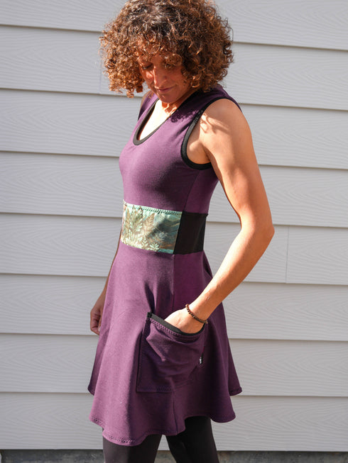 Woman wearing a purple dress with a belt standing against a gray wall.